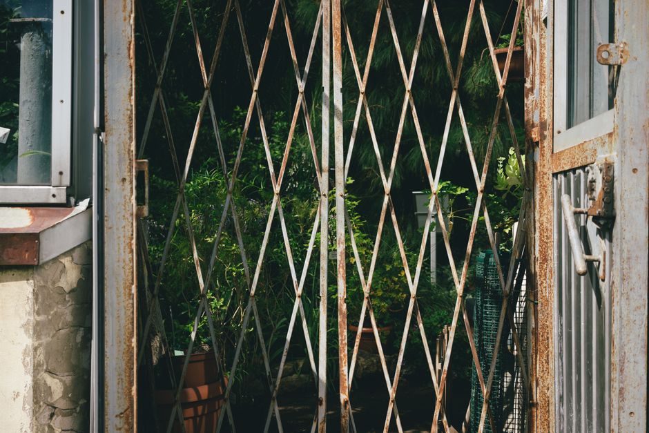 A weathered metal garden gate with a rusted, lattice-style grille partially open, revealing a lush, green garden beyond. The garden includes various plants with dense foliage, a flowering shrub, and potted greenery, all situated on a narrow pathway. To the left, a small section of stone and brickwork is visible, with a portion of a window frame. The lighting is natural, indicating daytime, and the scene emphasizes outdoor garden maintenance and landscaping work, aligning with the guides for narrow garden access by Gardeners Kennington. The overall composition highlights the textures of the rusted metal, lush plants, and uneven pathway, providing an accurate visual description suitable for outdoor garden content.