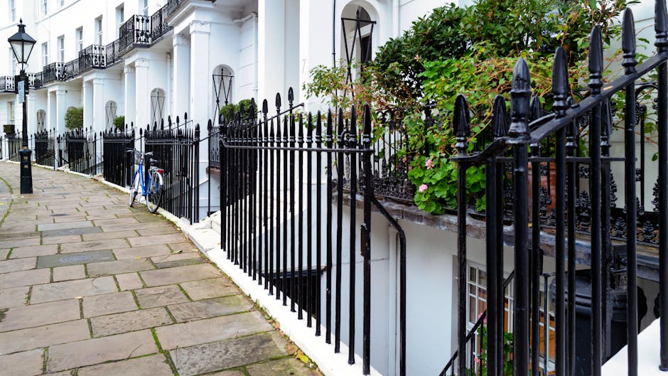 A paved walkway along a row of white terraced houses with black wrought iron railings. The pathway is composed of rectangular stone slabs with moss and slight discoloration on some edges, indicating outdoor use. Behind the railings, there are small garden beds with lush green foliage, including a climbing plant with pink flowers growing on a trellis or railing. The houses feature large windows with black metal frames and decorative ironwork beneath the windowsills. The scene is illuminated by natural daylight, creating a bright and clear outdoor setting. The overall atmosphere depicts a tidy, well-maintained residential street, with gardening and outdoor maintenance actions likely carried out by Gardeners Kennington, who specialize in garden care and landscaping work in the SE11 area.