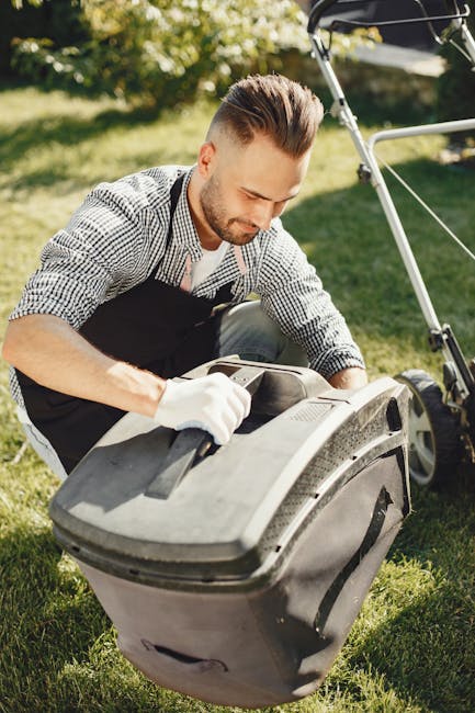 A man with a neatly styled haircut and a trimmed beard is engaged in garden maintenance, kneeling on a lush green lawn while opening a large black garden waste bin. He is dressed in a checked shirt, black apron, and white gloves, demonstrating careful handling. Surrounding him, the garden features a well-maintained lawn with freshly cut grass, a border of flowering plants, and a hedge in the background, indicating recent trimming. Sunlight illuminates the scene, casting soft shadows and highlighting the textures of the grass, soil, and the man's clothing. Nearby, a lawnmower with a silver handlebar is visible, suggesting ongoing landscaping work. This image effectively captures the outdoor setting of a typical backyard garden ready for routine care and tidy-up efforts, consistent with the garden maintenance and landscaping services offered by Gardeners Kennington. The peaceful environment and meticulous attention to detail reflect quality gardening practices suitable for a SE11 terraced garden, supporting homeowners and gardeners in maintaining a vibrant outdoor space.